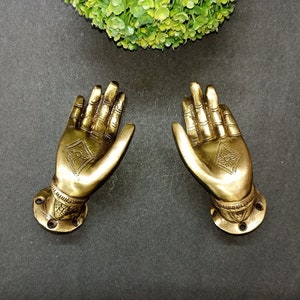 May include: A pair of gold-toned metal door handles shaped like open hands with intricate designs. The handles are mounted on round bases.
