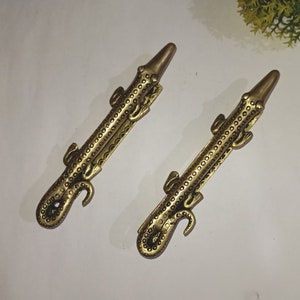 May include: Two gold-toned alligator-shaped door handles. The handles are detailed with a textured pattern of small circles.