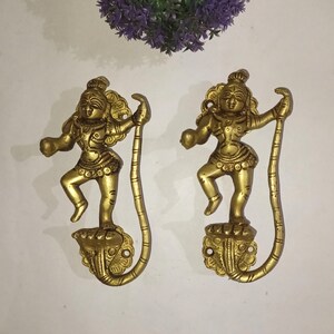 Könnte beinhalten: Zwei goldfarbene, dekorative Haken aus Metall in Form von tanzenden Figuren. Jede Figur hat einen langen, dünnen Schwanz, der sich zu einem Haken krümmt. Die Figuren stehen auf einem Sockel mit aufwendigen Details.