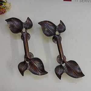 May include: Two ornate bronze door handles with a leaf design. Each handle features two leaves with detailed veins and a stem with a small hole in the center.