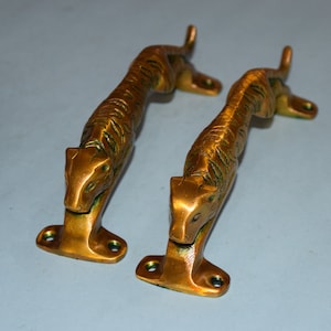 May include: Two brass tiger-shaped door handles. The handles are detailed with etched stripes and facial features. Each handle has a base with two screw holes for mounting. The handles are a warm gold color.