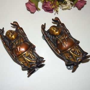 May include: Two gold and bronze coloured metal door handles shaped like a laughing Buddha figure. The handles are mounted on a black metal base.