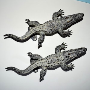 May include: Two decorative alligator figurines in a distressed, metallic finish. The alligators are detailed with textured scales and have mounting hardware attached. The color scheme is primarily gray and black, with hints of bronze.