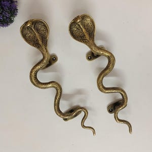 May include: Two brass-colored door handles shaped like cobras. The cobra heads are detailed with etched patterns, and the serpentine bodies curve gracefully. The handles are mounted on a white surface, suggesting a decorative or artistic theme.