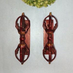 May include: Two ornate red and gold Tibetan prayer wheels with intricate designs. The wheels are mounted on wooden plaques.