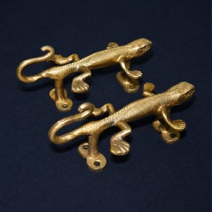 May include: Two gold-colored lizard-shaped door handles. Each handle features a detailed lizard design with textured skin and a curved tail. The handles are mounted on a base with screw holes for easy installation. The background is a dark blue.