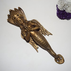 Może przedstawiać: Brązowa klamka do drzwi w kształcie mitycznej postaci z koroną, skrzydłami i ogonem syreny. Postać jest w pozycji modlitewnej. Klamka ma skomplikowane detale i dekoracyjne zakończenie.