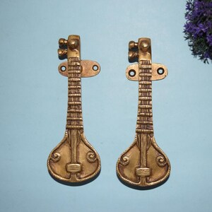 Op de afbeelding: Twee messingkleurige decoratieve deurklinken in de vorm van een snaarinstrument, waarschijnlijk een veena. De klinken hebben een gedetailleerd ontwerp, inclusief stemschroeven en een afgeronde body. Op een lichtblauwe achtergrond.