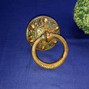 Könnte beinhalten: Verzierter Türklopfer aus Bronze mit einer kreisförmigen Grundplatte und einem Ringgriff. Die Grundplatte weist ein detailliertes Blumendesign mit einer Grünspan-Oberfläche auf. Der Ringgriff hat ein strukturiertes Muster.