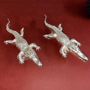 Peut inclure: Deux figurines d'alligator en argent avec des écailles détaillées et des gueules ouvertes. Les alligators sont posés la tête en avant et la queue enroulée.