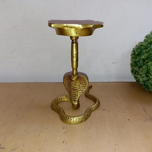 以下が含まれることがあります： 丸いトップを持つコブラの形をした金色の金属製の香炉。香炉は木製の表面の上に置かれています。