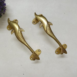 Könnte beinhalten: Zwei goldfarbene, delphin-förmige Türklinken mit strukturierter Oberfläche. Die Griffe haben ein schlichtes Design mit abgerundeter Oberseite und einem geschwungenen Schwanz.