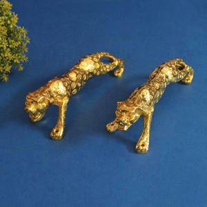 May include: Two golden cheetah figurines in a crawling pose. The cheetahs have detailed patterns and are set against a blue background. These decorative sculptures add a touch of elegance to any space.