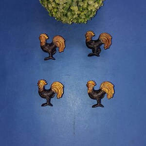 May include: Four small, decorative rooster figurines. Each rooster is black with gold-colored tail feathers and a comb. The figurines are arranged on a blue surface, with a green floral element in the upper corner.