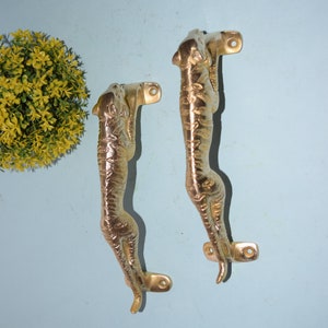Puede incluir: Dos tiradores de puerta de metal dorado con forma de tigre. Los tiradores están montados en una pared azul claro.
