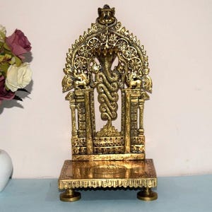 Pode incluir: Uma escultura dourada e ornamentada que apresenta uma cobra com um fundo arqueado. A escultura é montada sobre uma base retangular com quatro pequenos pés. O design inclui padrões e figuras intrincados.