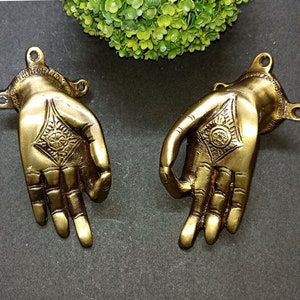 Peut inclure: Deux poignées de porte en métal de couleur dorée en forme de main avec des motifs complexes. Les poignées sont face à face et se trouvent sur un fond gris foncé.