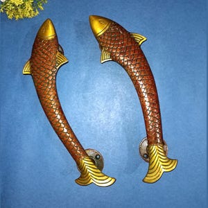 Op de afbeelding: Twee decoratieve deurklinken in de vorm van vissen. De handgrepen zijn bronskleurig met een schubbenpatroon en gouden accenten op de kop en staart. De achtergrond is blauw.