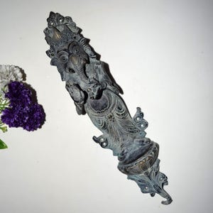 Könnte beinhalten: Eine detaillierte, antik anmutende Bronzestatue der hinduistischen Gottheit Ganesha. Die Statue ist länglich mit aufwendigen Schnitzereien und einem detaillierten Kopfschmuck. Die Figur wird mit einem Elefantenkopf und mehreren Armen dargestellt, die verschiedene Objekte halten. Die Statue ist an einer weißen Wand befestigt.