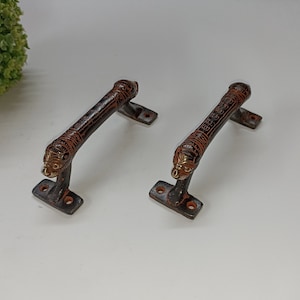 May include: Two antique-style door handles with ornate designs. The handles are made of dark metal with a copper finish and feature lion head details.