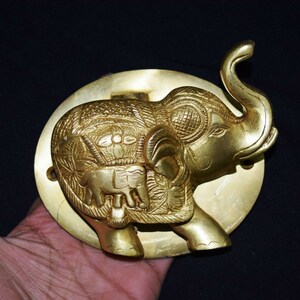 May include: A gold-colored brass door knocker shaped like an elephant with intricate details. The elephant has a raised trunk and is standing on a round base.