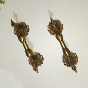 May include: A pair of antique brass door handles. Each handle has an ornate design with a central cylindrical grip and decorative floral elements. The handles are a warm, golden colour.
