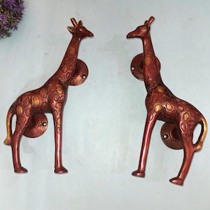 May include: Two decorative giraffe-shaped door pulls. The pulls are made of reddish-brown metal with gold-colored spots and accents. Each giraffe has a circular mounting plate. The door pulls are designed to be mounted on a door or cabinet.