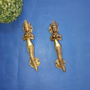 Puede incluir: Dos tiradores de puerta de latón con forma de sirena. Cada tirador presenta una figura detallada de sirena con las manos juntas en oración, una corona decorativa y una cola escamada. Los tiradores están montados sobre una base floral, sobre fondo azul.
