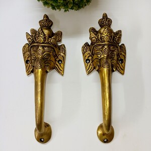 Op de afbeelding: Twee messingkleurige deurgrepen, elk met een olifantenkopontwerp met ingewikkelde details. De handgrepen hebben een gebogen vorm en zijn op een wit oppervlak gemonteerd. De handgrepen zijn decoratief en sierlijk.