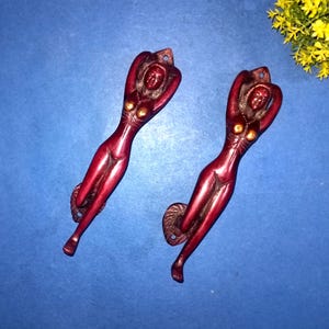 May include: Two red-brown metal door handles in the shape of a reclining woman. The handles have a detailed design, with the woman's arms raised above her head. The handles are on a blue background.