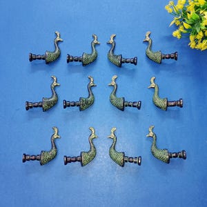 Puede incluir: Doce tiradores decorativos con forma de pavo real. Cada tirador presenta un cuerpo verde con detalles dorados y una base marrón oscuro. Están dispuestos sobre una superficie azul, con un pequeño ramo de flores amarillas en la esquina superior derecha.