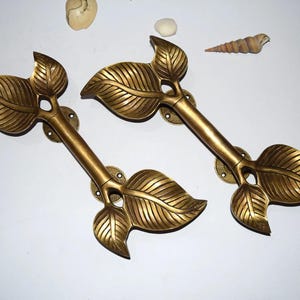 Puede incluir: Dos tiradores de puerta de latón con forma de hoja. Cada tirador presenta un tallo central con tres hojas detalladas. Los tiradores miden aproximadamente 30 cm de largo y están montados sobre placas circulares con orificios para tornillos. Los tiradores son de color dorado cálido.