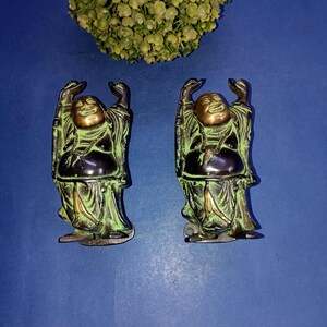 以下が含まれることがあります： 青い表面の上に立っている、緑と金のブロンズ製の笑う仏陀の小さな像が2つ。それぞれの像は、祝福のしぐさで腕を上げている。