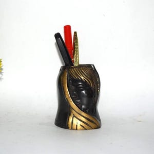 May include: A black and gold pen holder with a carved face design. The pen holder contains a black pen, a red pen, and a gold pen. The pen holder is made of a dark material and has gold accents.