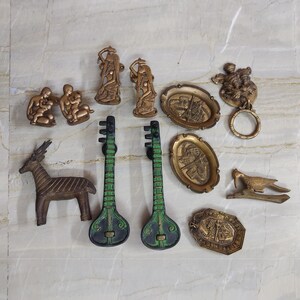 May include: A collection of assorted brass decorative items including two green and black stringed instrument handles, a bronze deer, two kneeling figures, two oval plaques with city scenes, a bird on a branch, and a circular door knocker.