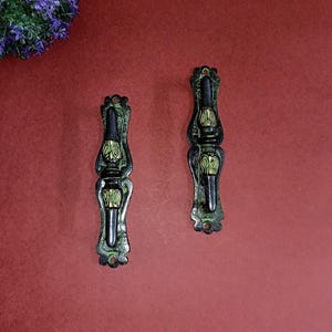 May include: Two black and green metal drawer pulls with ornate designs. The pulls have a decorative, floral pattern.