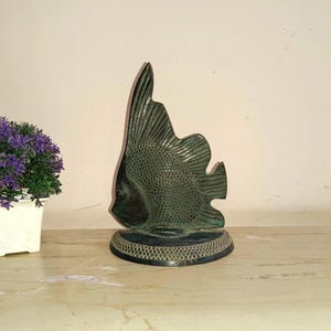 May include: A bronze-colored fish sculpture with detailed fins and scales. The fish is mounted on a decorative base with a patterned border. The sculpture is a decorative item, suitable for home decor.