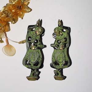 May include: Two green and gold metal door handles shaped like dancing women. The handles are detailed with intricate designs and have a traditional Indian style.