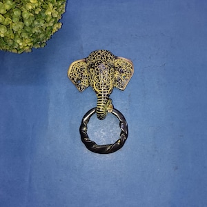 May include: A brass elephant head door knocker with a textured design and a dark metal ring handle. The elephant's head is detailed with etched patterns and has large, flared ears. The knocker is set against a blue background.