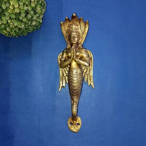 Puede incluir: Manija de puerta de latón con una escultura detallada de una deidad con capucha de cobra, manos en oración y cuerpo inferior escamoso. La manija tiene un acabado dorado y está montada sobre una superficie azul. La manija mide aproximadamente 25 cm de largo.