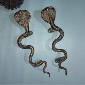 May include: A pair of metal door handles shaped like cobras with an antique bronze finish. Each handle features intricate engraved patterns along the body and hood of the snake, with two mounting points for attachment to a door surface.