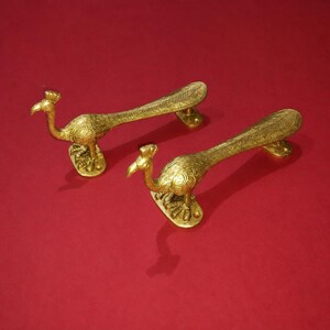 Puede incluir: Dos tiradores de puerta dorados con forma de pavo real. Los tiradores tienen tallas detalladas del cuerpo y las plumas de la cola del ave. Se encuentran sobre un fondo rojo.