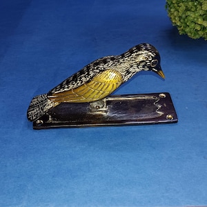 Puede incluir: Un llamador de puerta de latón con forma de pájaro con alas negras y amarillas. El pájaro está posado sobre una base rectangular.