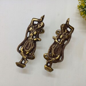 Puede incluir: Dos manijas de puerta antiguas de latón con forma de figuras danzantes. Las manijas están detalladas con diseños intrincados y tienen un color marrón dorado cálido. Se montan sobre una superficie blanca, con un pequeño arreglo floral amarillo en el fondo.