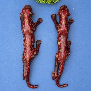 May include: Two red leopard-shaped wall hooks with gold-colored spots and a textured finish. The hooks are positioned vertically against a blue background, with a green plant in the upper portion of the image.