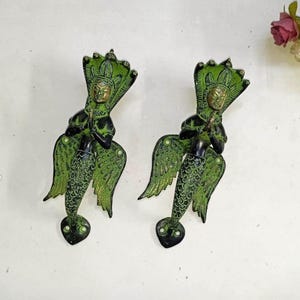Pode incluir: Duas puxadores de porta antigos de bronze com sereias. Cada puxador apresenta uma sereia com um corpo verde texturizado, braços pretos e um adorno de cabeça detalhado. As sereias têm cauda de peixe e seguram um instrumento musical. Os puxadores têm cerca de 15 cm de altura.