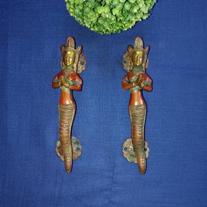 以下が含まれることがあります： 祈りを捧げる女性の形をした、装飾的な真鍮製のドアノブのペア。ノブは金色と赤色のトーンが混ざり合っています。