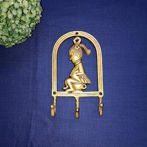 May include: A gold-colored wall hook featuring a decorative design of a seated figure within an arched frame. The hook has three lower hooks for hanging items. The design is set against a dark blue background.