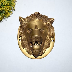 Puede incluir: Un abridor de botellas de cabeza de león dorado con un diseño detallado. La boca del león está abierta y los dientes son visibles. El abridor de botellas está hecho de latón y tiene una forma circular.