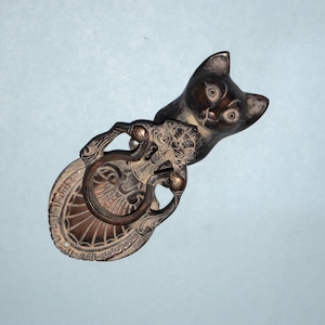 Puede incluir: Un tirador de cajón en forma de gato en tono bronce con un asa ovalada. La cabeza del gato está en la parte superior, con ojos grandes y una cara detallada. El asa presenta un diseño ornamentado con un tirador circular.